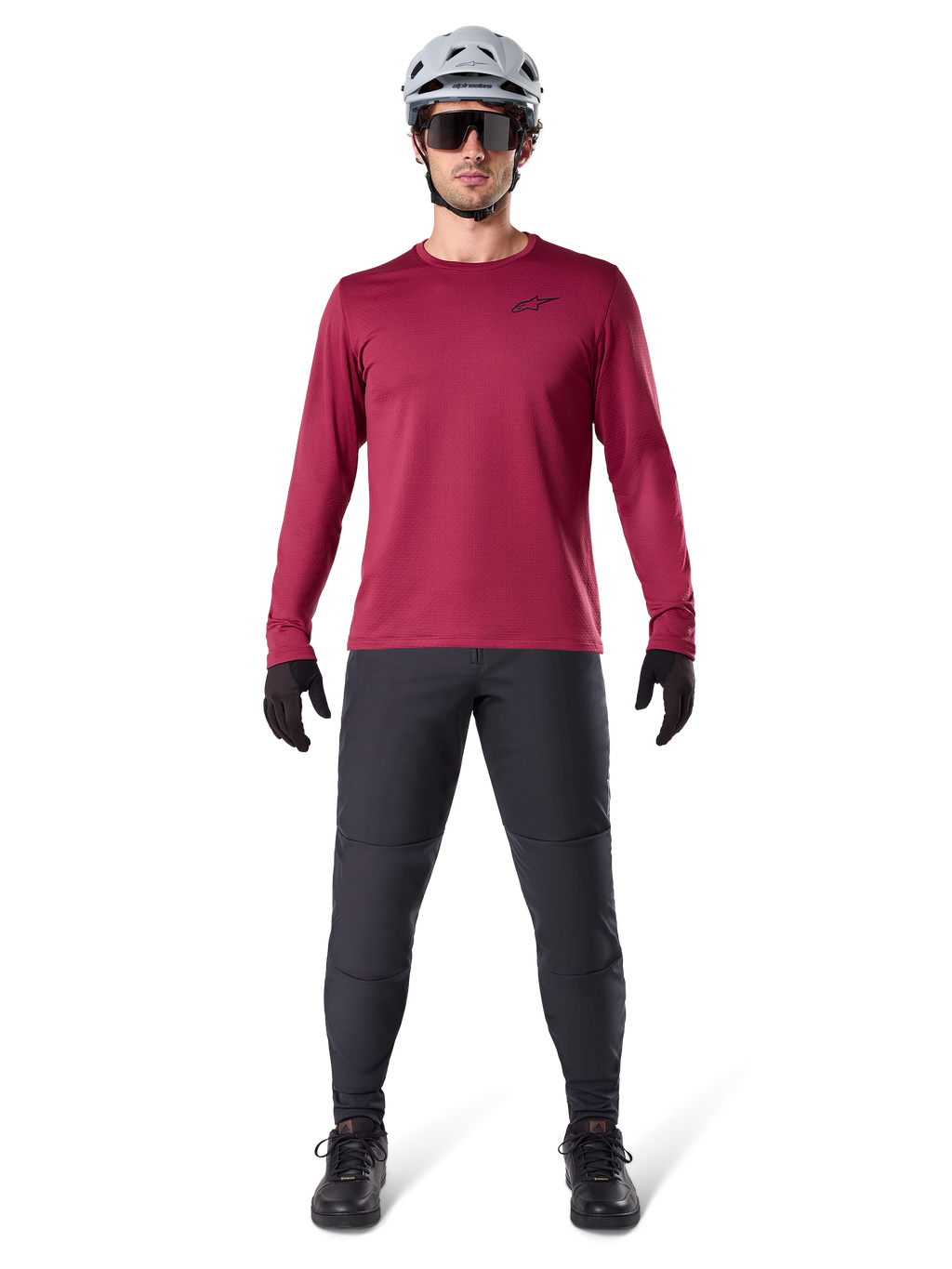 Męski model w koszulce termicznej Alpinestars A-Dura Thermal w kolorze bordowym, koszulka rowerowa z długim rękawem i izolacją, ciemnoszare spodnie MTB, biały kask, czarne okulary przeciwsłoneczne i czarne rękawiczki, profesjonalna odzież MTB na chłodne dni