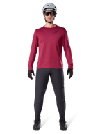 Męski model w koszulce termicznej Alpinestars A-Dura Thermal w kolorze bordowym, koszulka rowerowa z długim rękawem i izolacją, ciemnoszare spodnie MTB, biały kask, czarne okulary przeciwsłoneczne i czarne rękawiczki, profesjonalna odzież MTB na chłodne dni