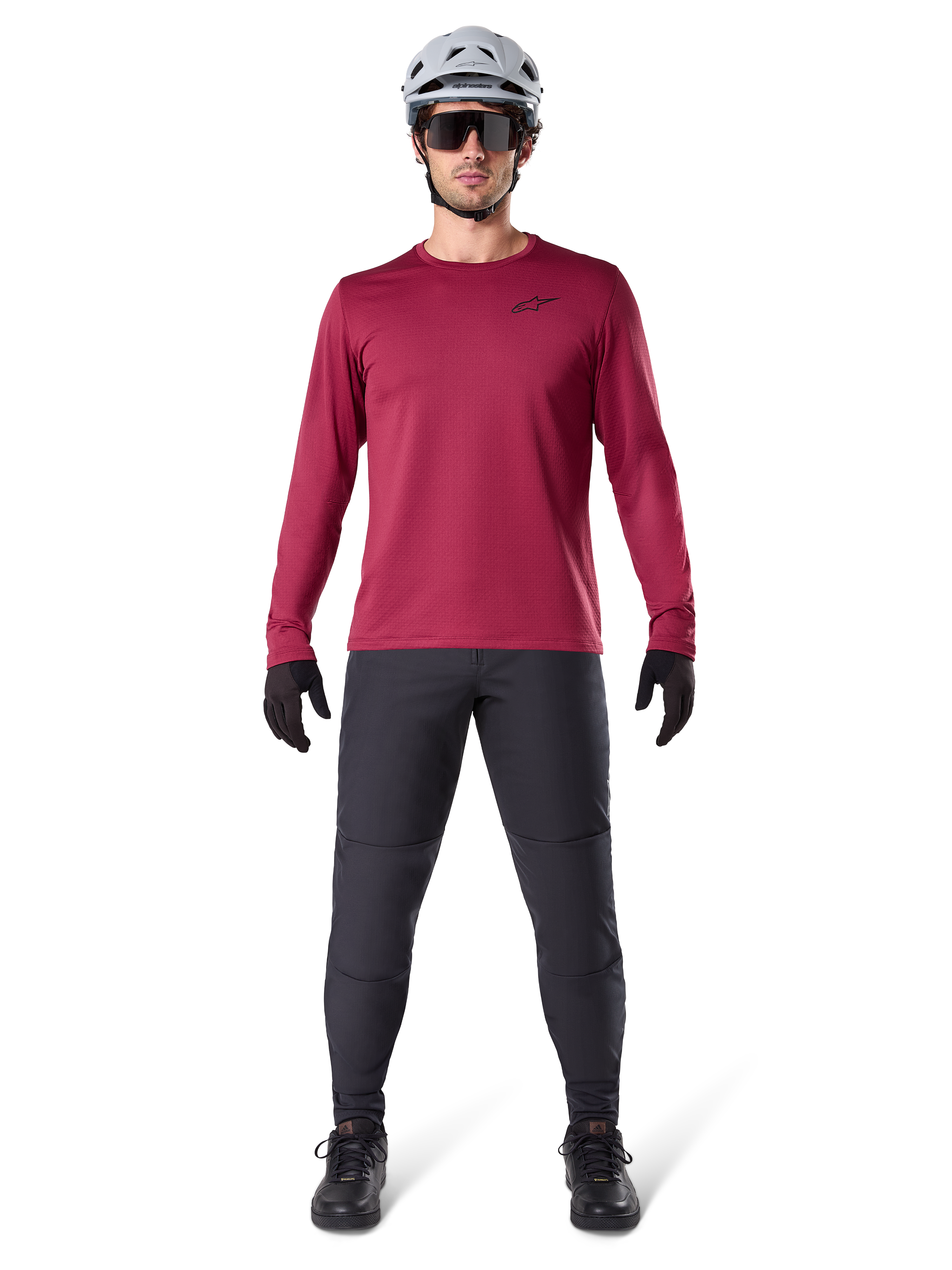 Męski model w koszulce termicznej Alpinestars A-Dura Thermal w kolorze bordowym, koszulka rowerowa z długim rękawem i izolacją, ciemnoszare spodnie MTB, biały kask, czarne okulary przeciwsłoneczne i czarne rękawiczki, profesjonalna odzież MTB na chłodne dni