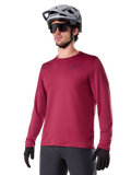 Alpinestars A-Dura Thermal Jersey, koszulka rowerowa MTB z długim rękawem, bordowa, z izolującą podszewką z polaru i małym czarnym logo na klatce piersiowej, noszona z szarym kaskiem, czarnymi okularami przeciwsłonecznymi i czarnymi rękawiczkami, zaprojektowana do jazdy w chłodne dni