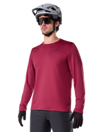 Alpinestars A-Dura Thermal Jersey, koszulka rowerowa MTB z długim rękawem, bordowa, z izolującą podszewką z polaru i małym czarnym logo na klatce piersiowej, noszona z szarym kaskiem, czarnymi okularami przeciwsłonecznymi i czarnymi rękawiczkami, zaprojektowana do jazdy w chłodne dni