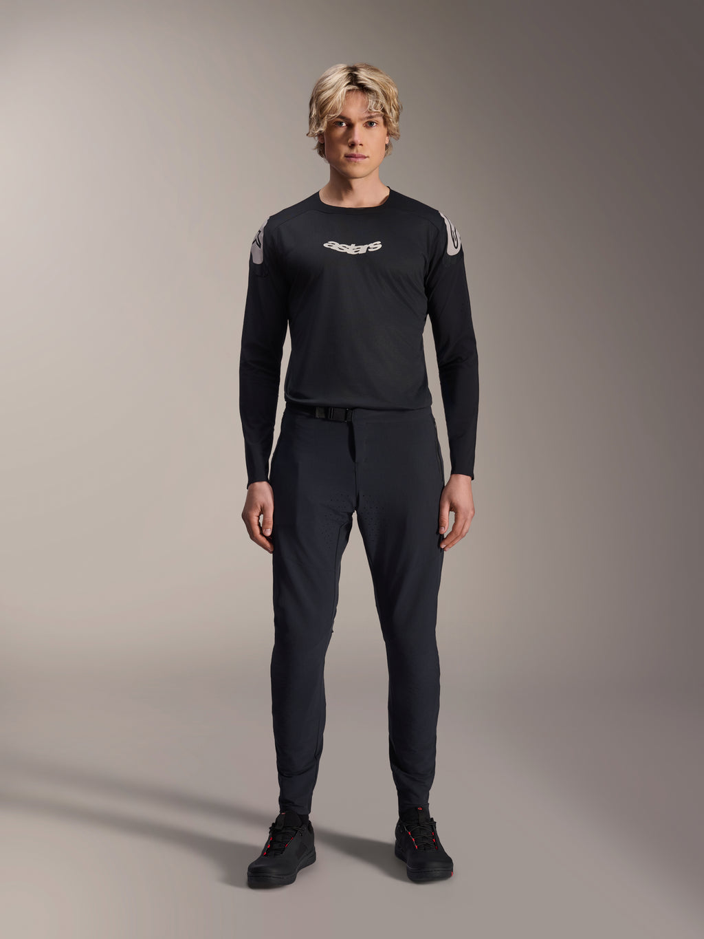 Spodnie Alpinestars A-Aria Elite, spodnie MTB w kolorze Black Titanium, prezentowane przez modela w dopasowanej czarnej koszulce z długim rękawem i sneakersach, wyczynowy krój slim-fit z magnetycznym zapięciem na klamrę, techniczna odzież do kolarstwa górskiego