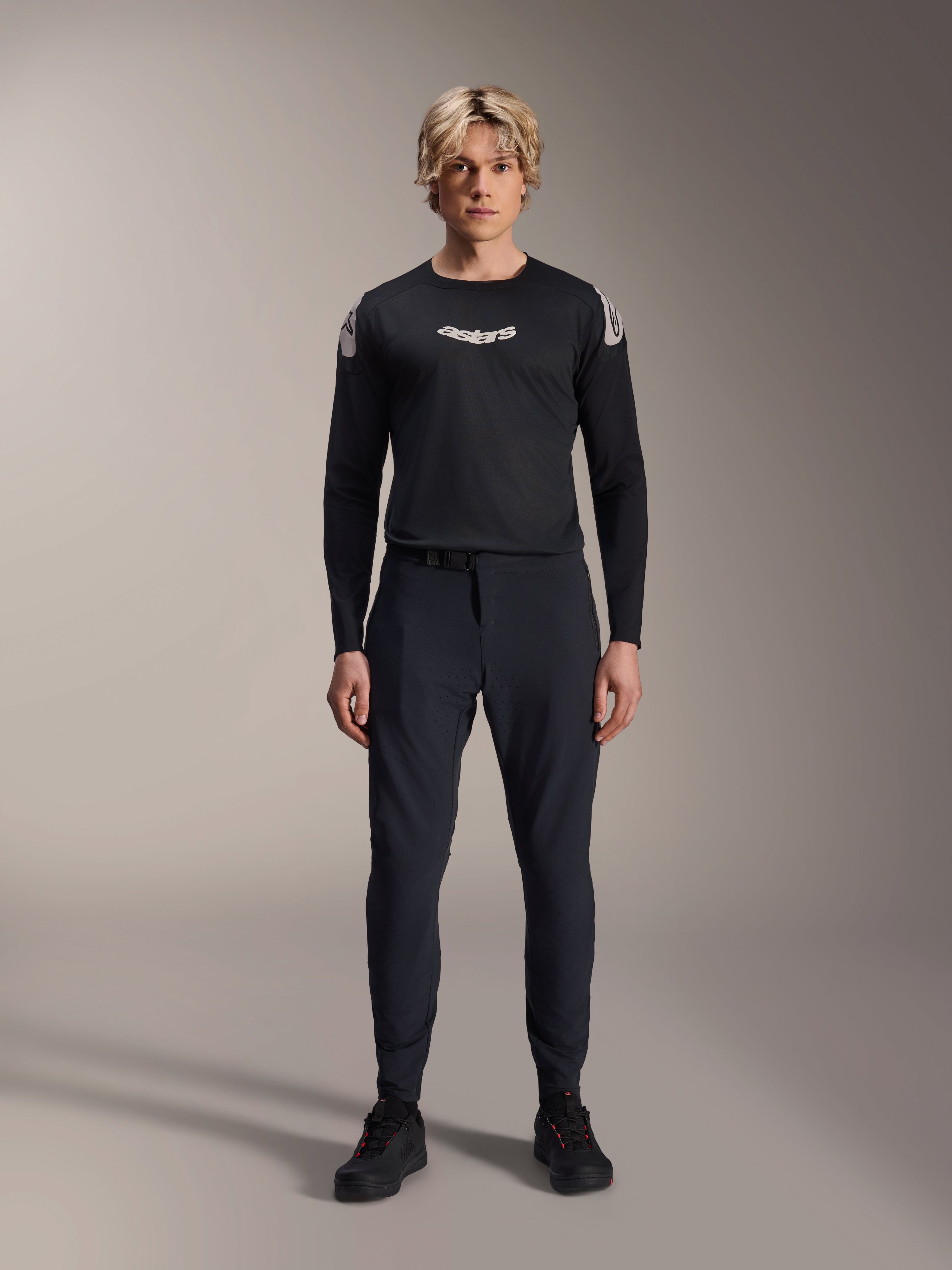 Spodnie Alpinestars A-Aria Elite, spodnie MTB w kolorze Black Titanium, prezentowane przez modela w dopasowanej czarnej koszulce z długim rękawem i sneakersach, wyczynowy krój slim-fit z magnetycznym zapięciem na klamrę, techniczna odzież do kolarstwa górskiego