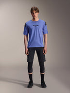 Male model wearing Alpinestars A-Aria Polartec Elite Short Sleeve Jersey in Baja Blue, black astars logo on the chest, paired with black MTB shorts, knee guards, and black cycling shoes, studio shot for mountain bike apparel
