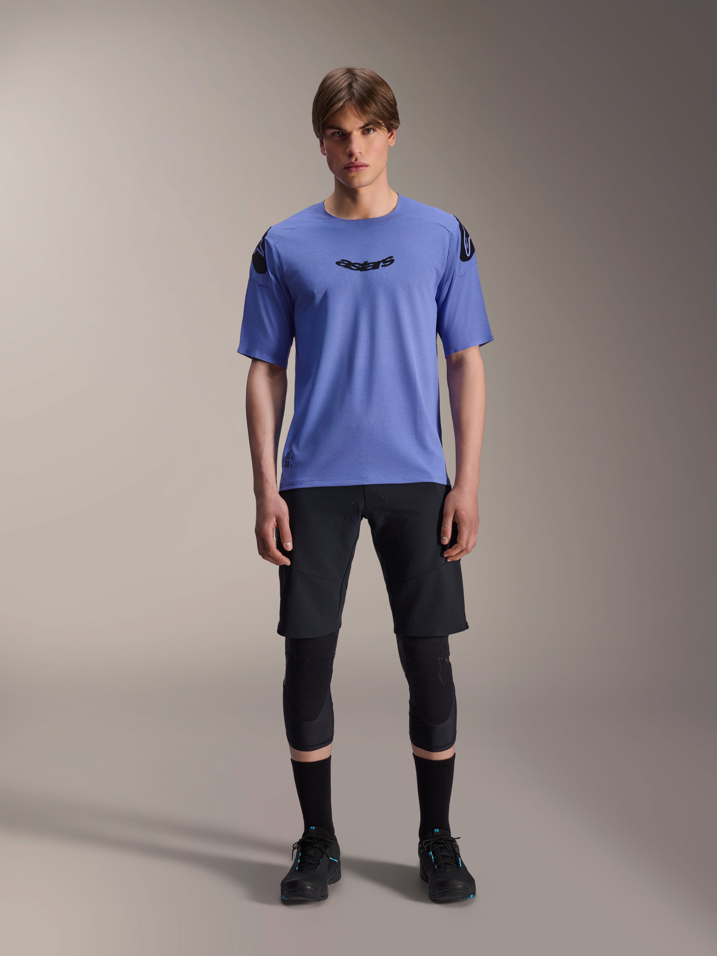 Male model wearing Alpinestars A-Aria Polartec Elite Short Sleeve Jersey in Baja Blue, black astars logo on the chest, paired with black MTB shorts, knee guards, and black cycling shoes, studio shot for mountain bike apparel