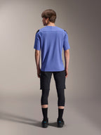 Mountain bike rider from behind wearing Alpinestars A-Aria Lite Shorts in black, paired with a blue short-sleeve jersey, black knee guards, and black socks, performance cycling gear for off-road trails