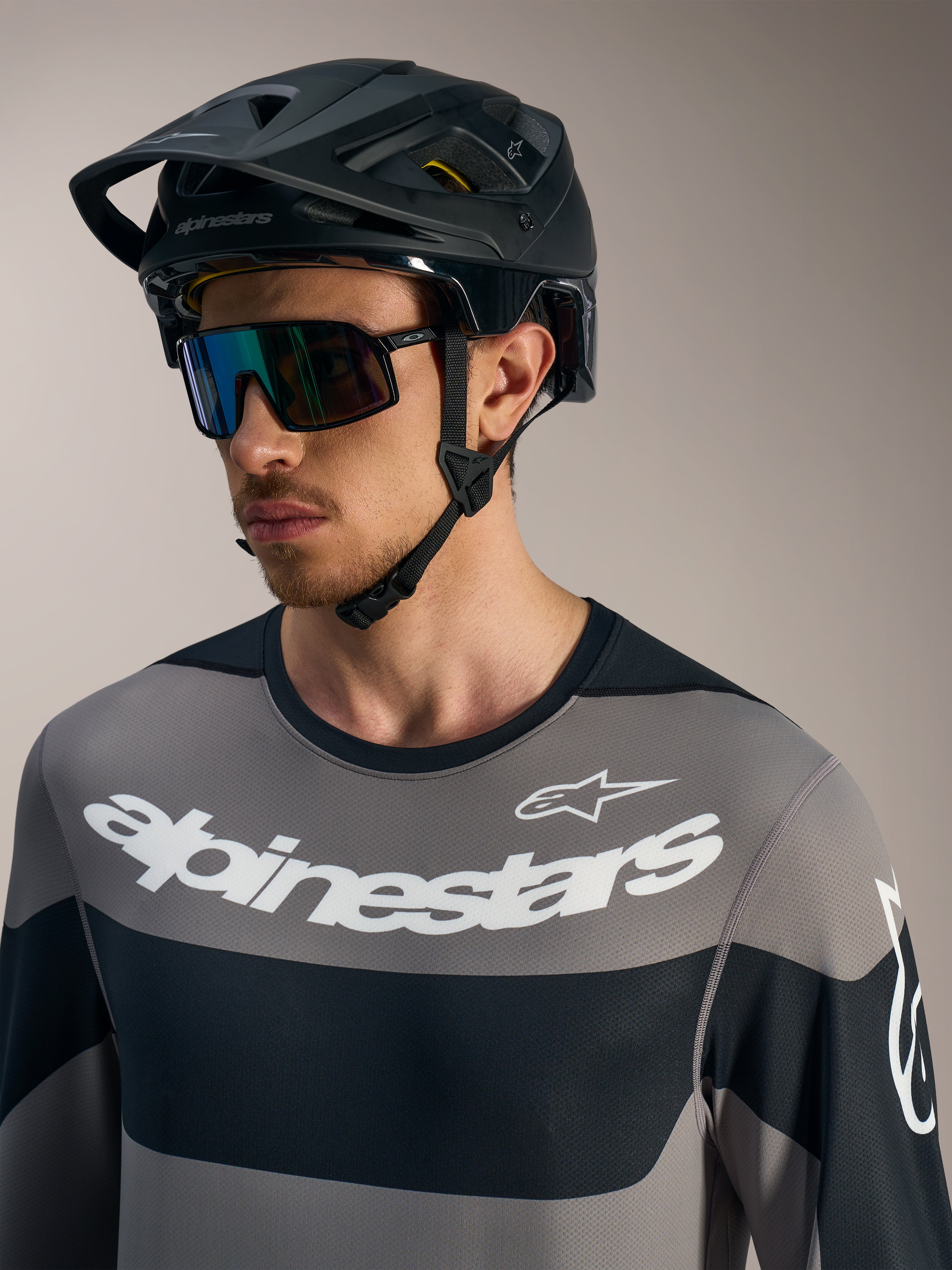 Alpinestars A-Dura Race LS, szara koszulka z białym logo, czarny kask MTB i okulary przeciwsłoneczne.