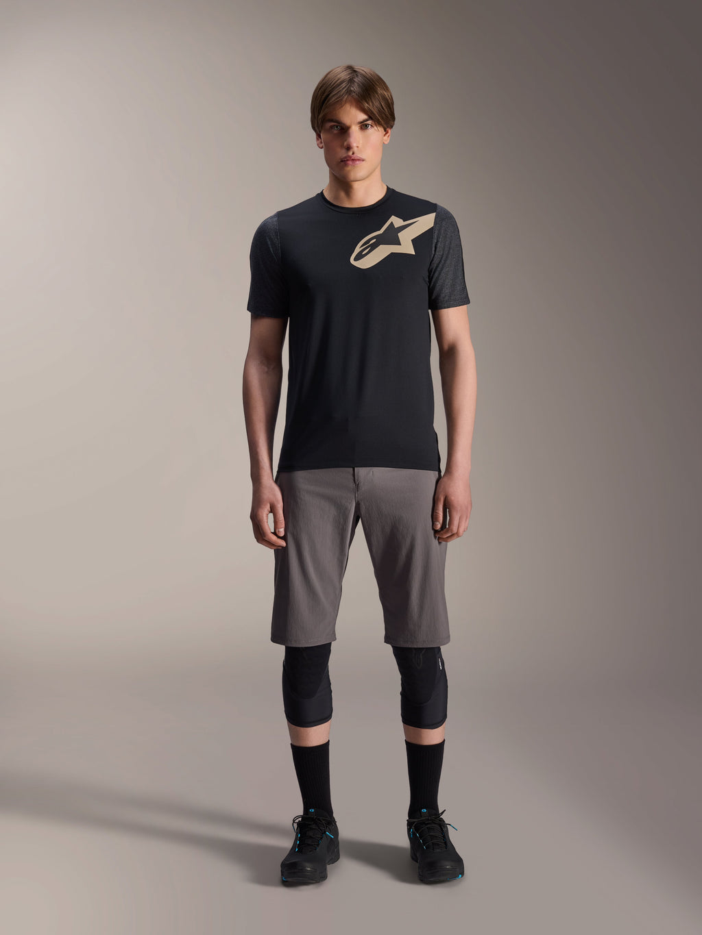Koszulka z krótkim rękawem Alpinestars A-Dura Lipan, czarna z rękawami w kolorze szary melanż i beżowym logo Astars na klatce piersiowej, prezentowana na modelu z szarymi szortami MTB i czarnymi ochraniaczami na kolana, profesjonalna odzież do jazdy MTB.