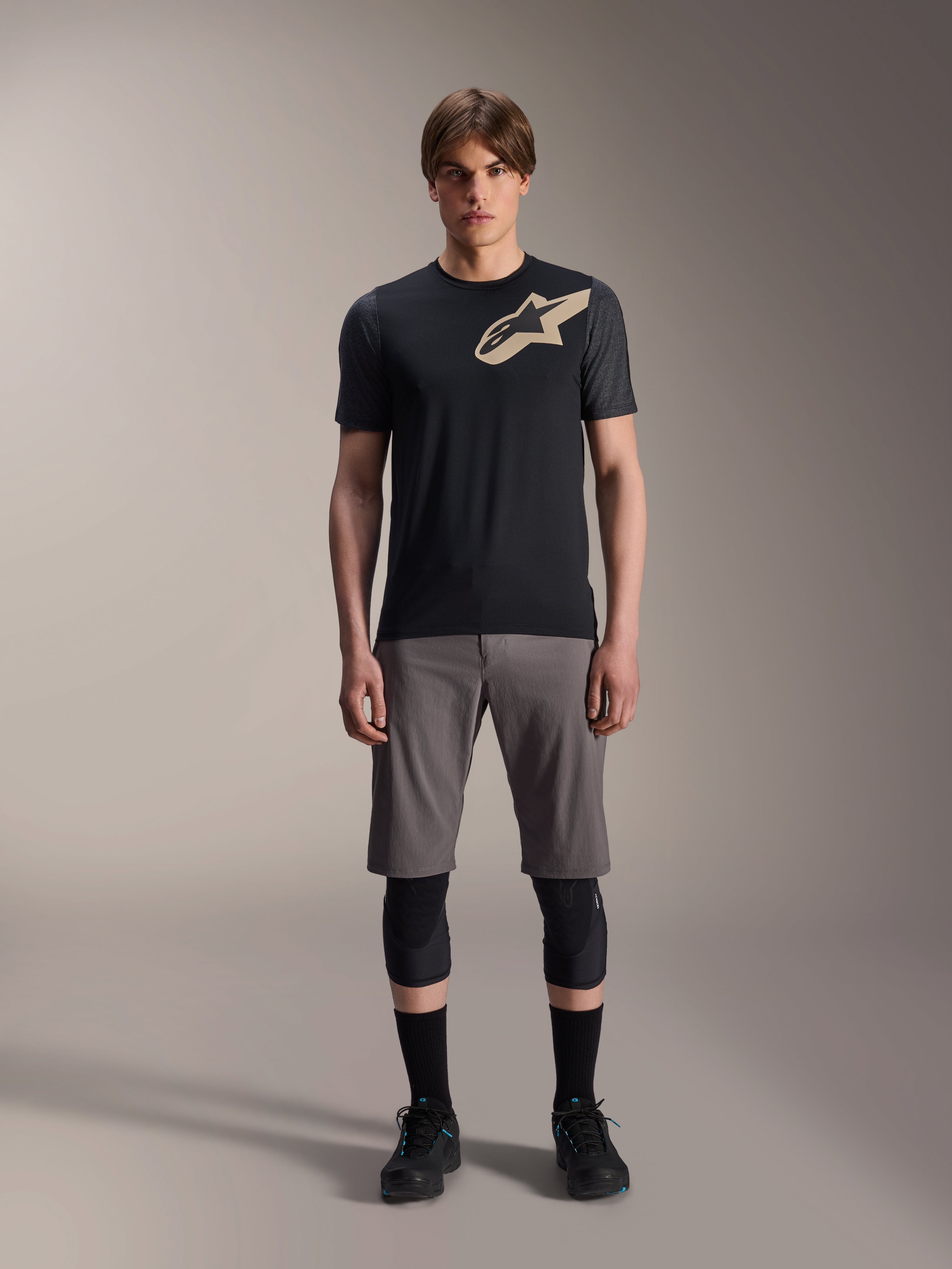 Koszulka z krótkim rękawem Alpinestars A-Dura Lipan, czarna z rękawami w kolorze szary melanż i beżowym logo Astars na klatce piersiowej, prezentowana na modelu z szarymi szortami MTB i czarnymi ochraniaczami na kolana, profesjonalna odzież do jazdy MTB.