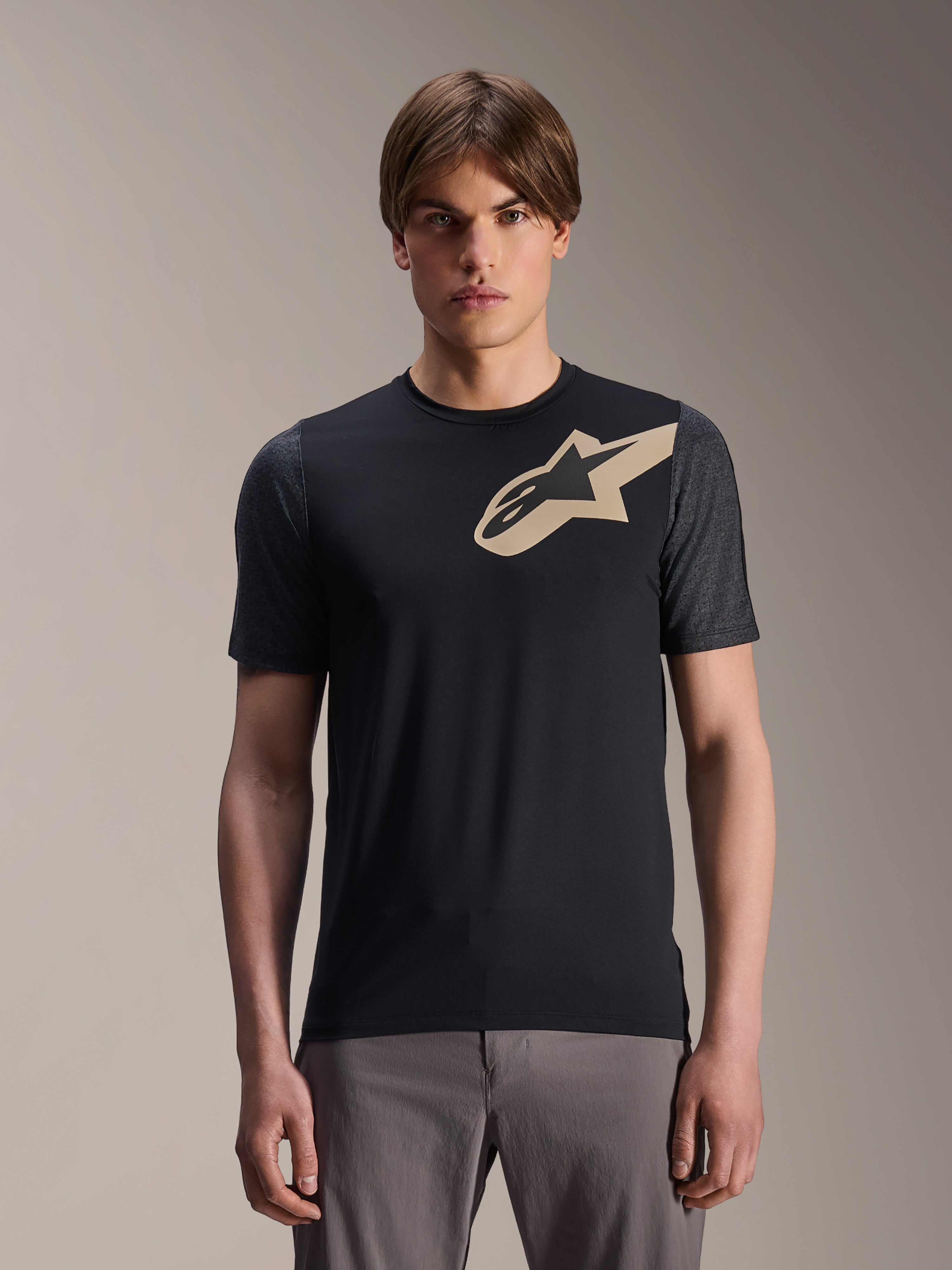 Męski model w koszulce Alpinestars A-Dura Lipan z krótkim rękawem, czarna koszulka rowerowa MTB z beżowym logo gwiazdy i ciemnoszarymi rękawami, w zestawie z szarymi spodniami rowerowymi, casualowa odzież funkcyjna