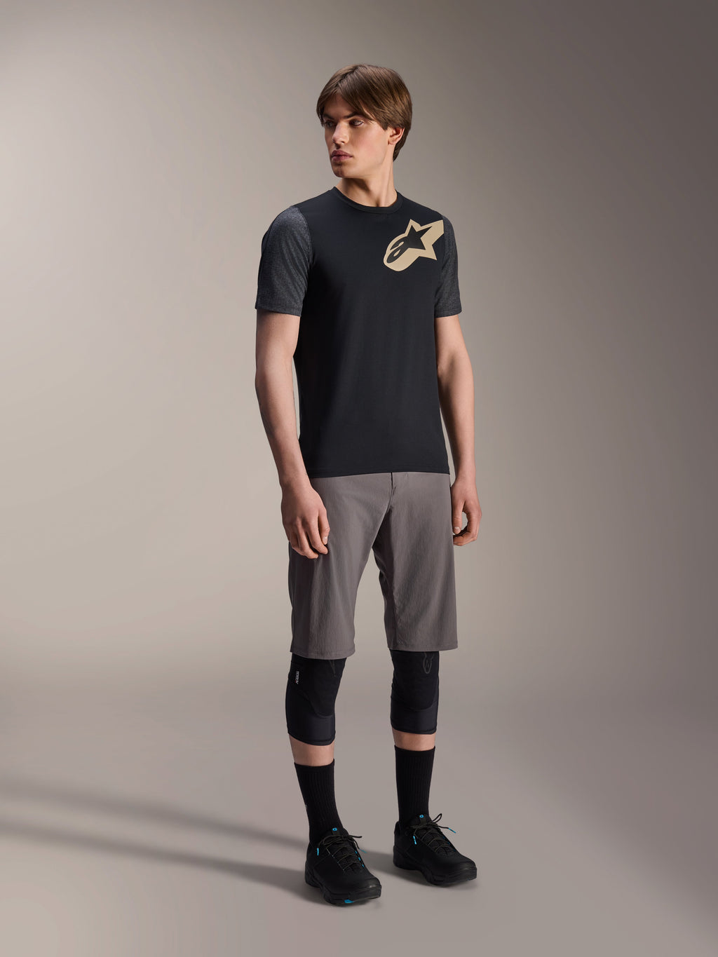 Męski model w koszulce Alpinestars A-Dura Lipan z krótkim rękawem w kolorze czarnym, z ciemnoszarymi rękawami i beżowym logo Astars na piersi, w zestawie z szarymi szortami rowerowymi, czarnymi ochraniaczami na kolana i czarnymi butami, kompletny strój do jazdy na rowerze górskim