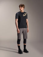 Męski model w koszulce Alpinestars A-Dura Lipan z krótkim rękawem w kolorze czarnym, z ciemnoszarymi rękawami i beżowym logo Astars na piersi, w zestawie z szarymi szortami rowerowymi, czarnymi ochraniaczami na kolana i czarnymi butami, kompletny strój do jazdy na rowerze górskim