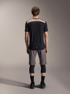 Alpinestars A-Dura Lipan Jersey - Short Sleeve, czarna z beżowym panelem na górze pleców, widok z tyłu pokazujący przedłużony tył, noszona z szarymi szortami MTB i czarnymi ochraniaczami kolan, wydajna odzież do jazdy na rowerze górskim