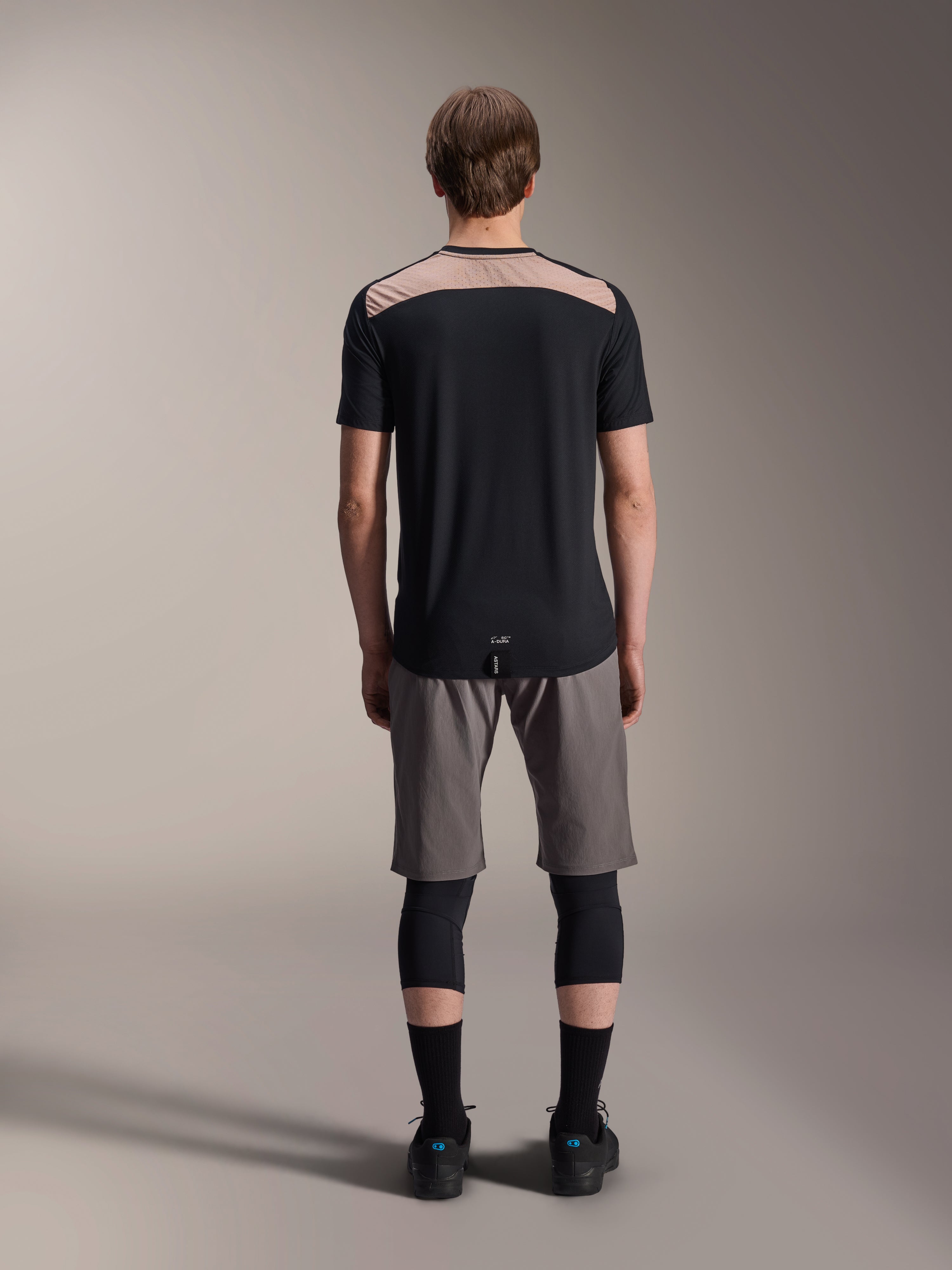 Alpinestars A-Dura Lipan Jersey - Short Sleeve, czarna z beżowym panelem na górze pleców, widok z tyłu pokazujący przedłużony tył, noszona z szarymi szortami MTB i czarnymi ochraniaczami kolan, wydajna odzież do jazdy na rowerze górskim