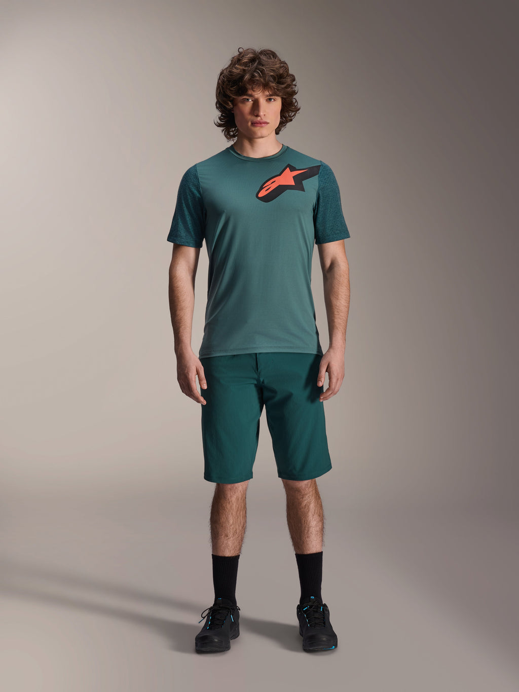 Alpinestars A-Dura Lipan Jersey, koszulka MTB z krótkim rękawem, zielona w odcieniu Silver Pine z czarno-pomarańczowym logo Astars na klatce piersiowej, w zestawie z dopasowanymi szortami i czarnym obuwiem, techniczna odzież do kolarstwa górskiego