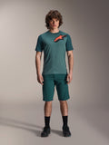 Alpinestars A-Dura Shorts w kolorze Dark Pine, męskie spodenki do kolarstwa górskiego, widoczne na modelu z dopasowaną turkusową koszulką z krótkim rękawem i czerwono-czarnym logo gwiazdy, czarne skarpetki i buty rowerowe, casualowa sportowa odzież MTB