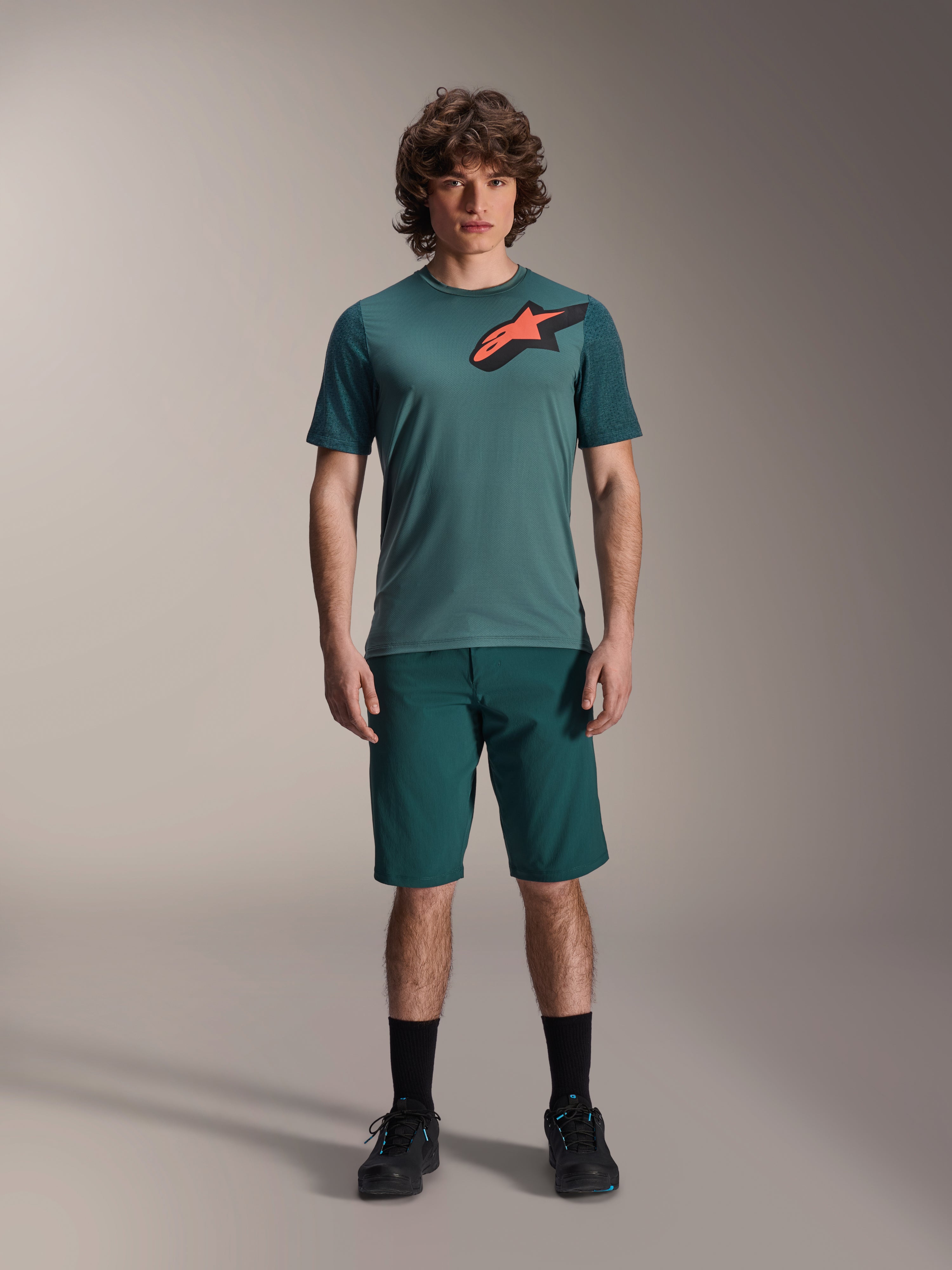 Alpinestars A-Dura Shorts w kolorze Dark Pine, męskie spodenki do kolarstwa górskiego, widoczne na modelu z dopasowaną turkusową koszulką z krótkim rękawem i czerwono-czarnym logo gwiazdy, czarne skarpetki i buty rowerowe, casualowa sportowa odzież MTB