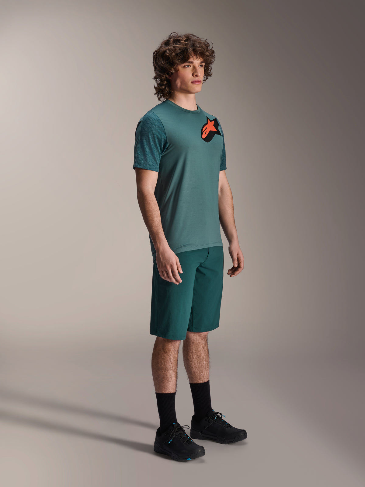 Koszulka Alpinestars A-Dura Lipan SS i spodenki A-Dura, strój rowerowy MTB, kolor Dark Pine, koszulka z dużym pomarańczowym logo gwiazdy, zestawiona z dopasowanymi spodenkami i czarnym obuwiem, profesjonalna odzież do kolarstwa górskiego
