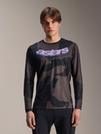 Alpinestars A-Dura Hollow Camo Long Sleeve Jersey, koszulka rowerowa MTB, czarno-szary wzór moro, fioletowa grafika „astars” na klatce piersiowej, prezentowana przez modela w czarnych spodniach, casualowy design o wysokiej wydajności