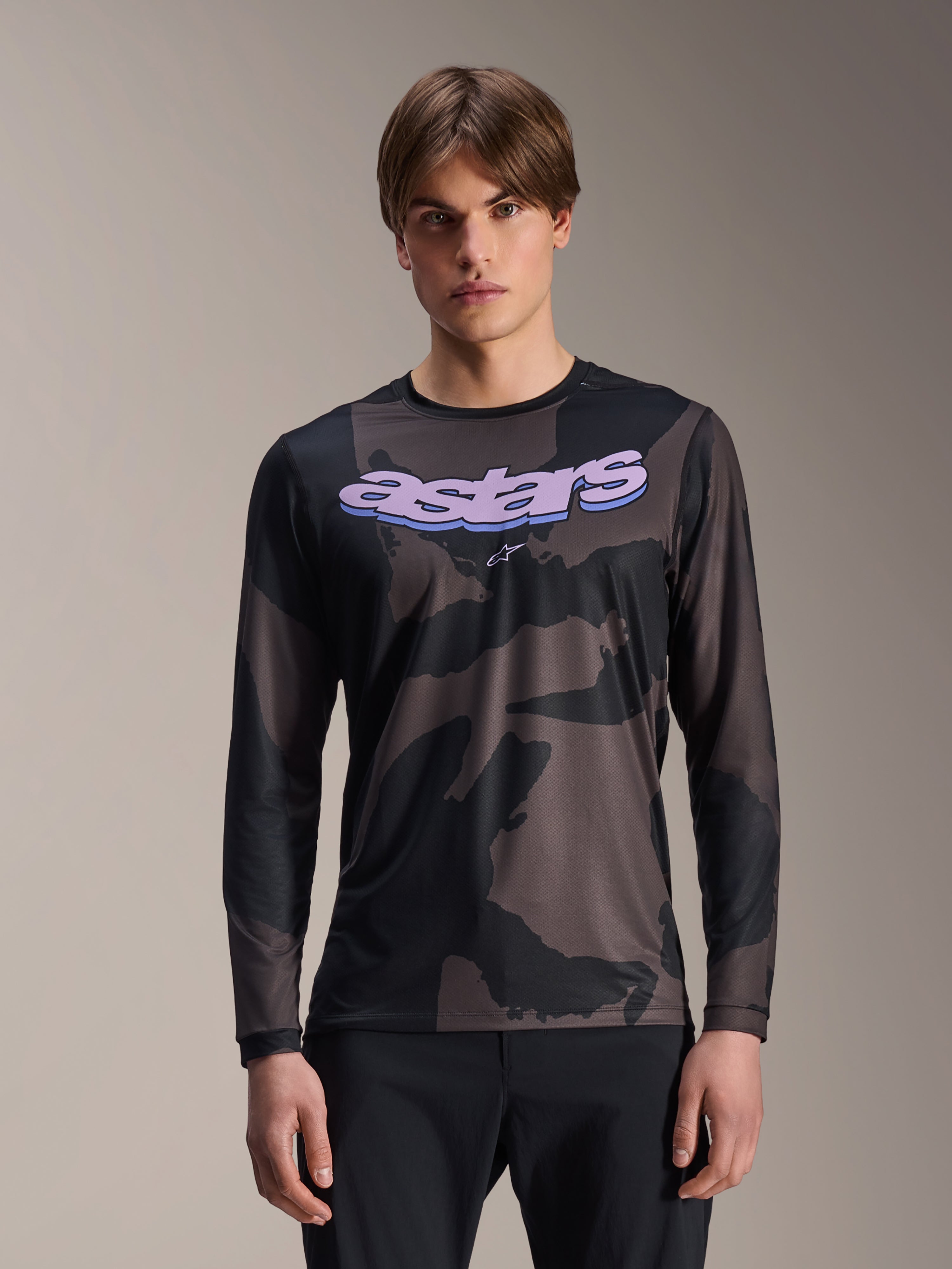Alpinestars A-Dura Hollow Camo Long Sleeve Jersey, koszulka rowerowa MTB, czarno-szary wzór moro, fioletowa grafika „astars” na klatce piersiowej, prezentowana przez modela w czarnych spodniach, casualowy design o wysokiej wydajności