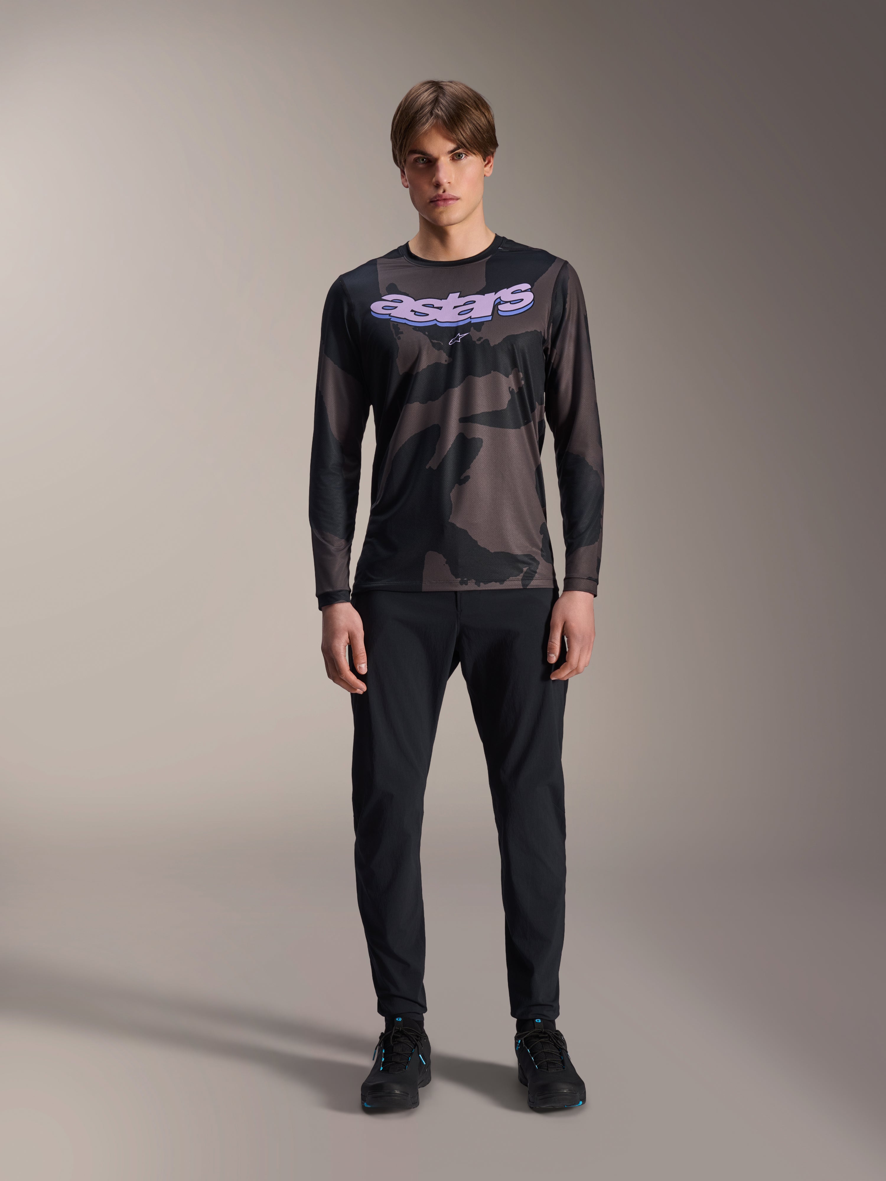 Model ubrany w koszulkę z długim rękawem Alpinestars A-Dura Hollow Camo w kolorze Black Camo, z fioletowym logo „astars” na klatce piersiowej, zestawioną z czarnymi spodniami i czarnymi butami rowerowymi, odzież do jazdy na rowerze górskim