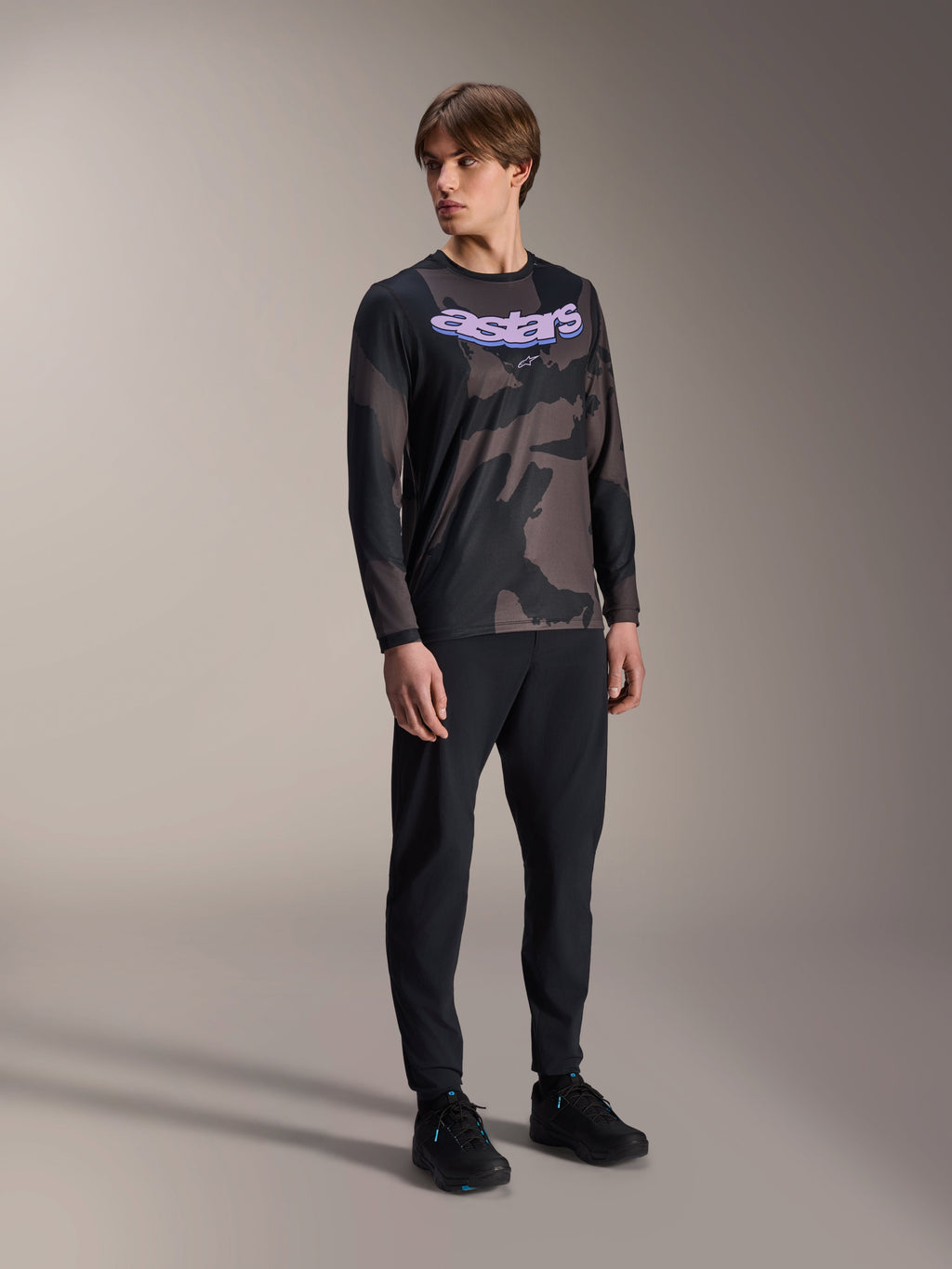 Męski model ubrany w koszulkę Alpinestars A-Dura Hollow Camo z długim rękawem, wzór Black Camo z fioletową grafiką 'astars' na piersi, czarne spodnie techniczne i czarne buty, casualowa odzież MTB