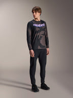 Męski model ubrany w koszulkę Alpinestars A-Dura Hollow Camo z długim rękawem, wzór Black Camo z fioletową grafiką 'astars' na piersi, czarne spodnie techniczne i czarne buty, casualowa odzież MTB