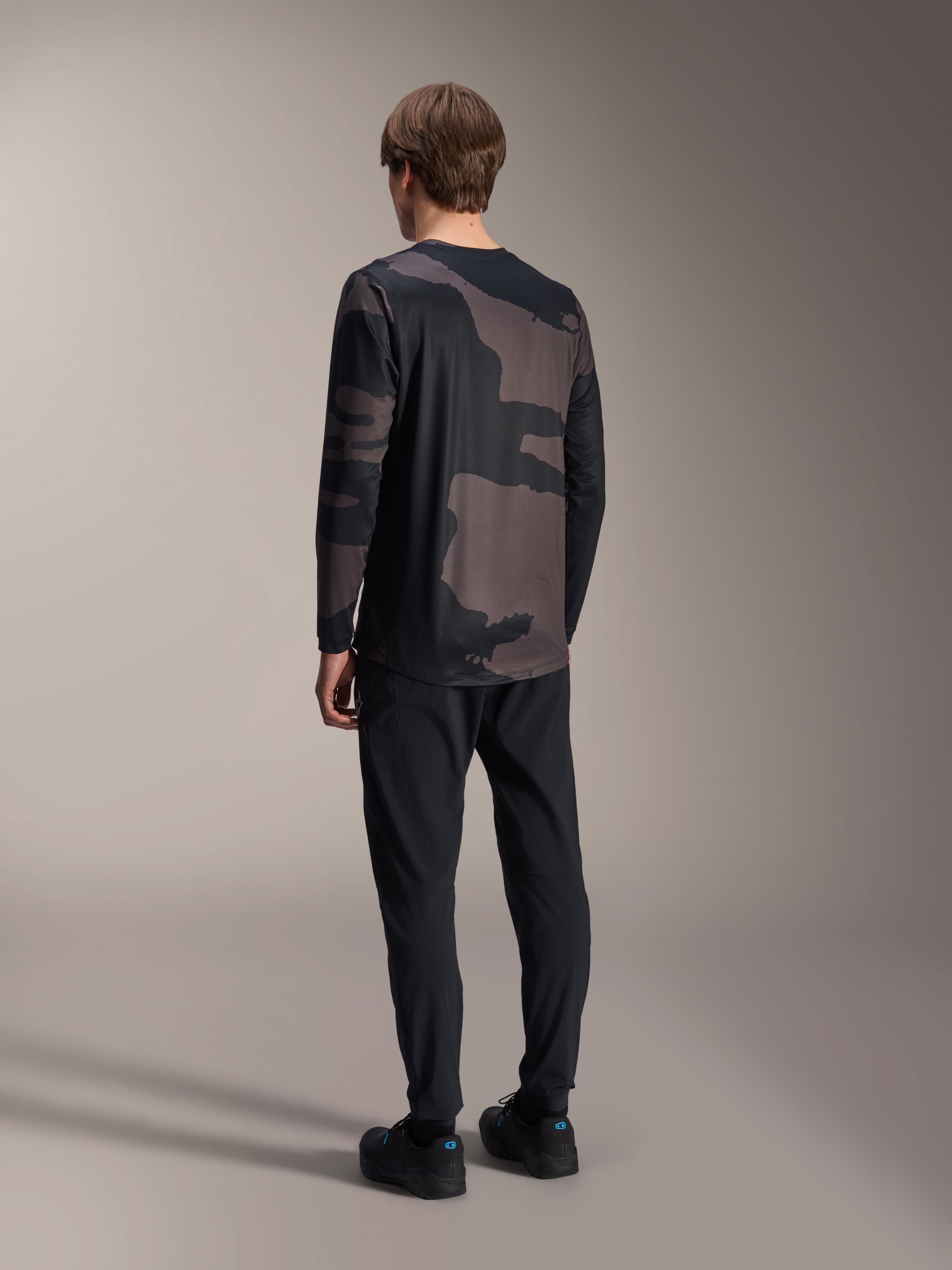 Alpinestars A-Dura Hollow Camo Long Sleeve Jersey, Black Camo, widok modelu z tyłu, techniczna koszulka rowerowa MTB z przedłużonym tyłem, zestawiona z czarnymi spodniami i butami rowerowymi, funkcjonalna odzież terenowa o swobodnym stylu