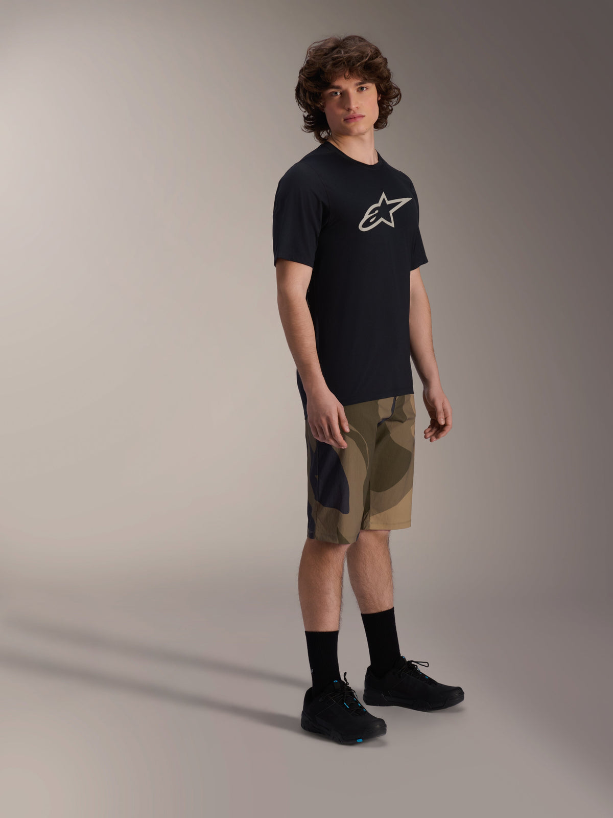 Alpinestars A-Dura Astar Short Sleeve Jersey, czarna koszulka z krótkim rękawem z białym logo na klatce piersiowej, szorty w kamuflażu i czarne buty MTB, casualowa odzież do jazdy na rowerze górskim