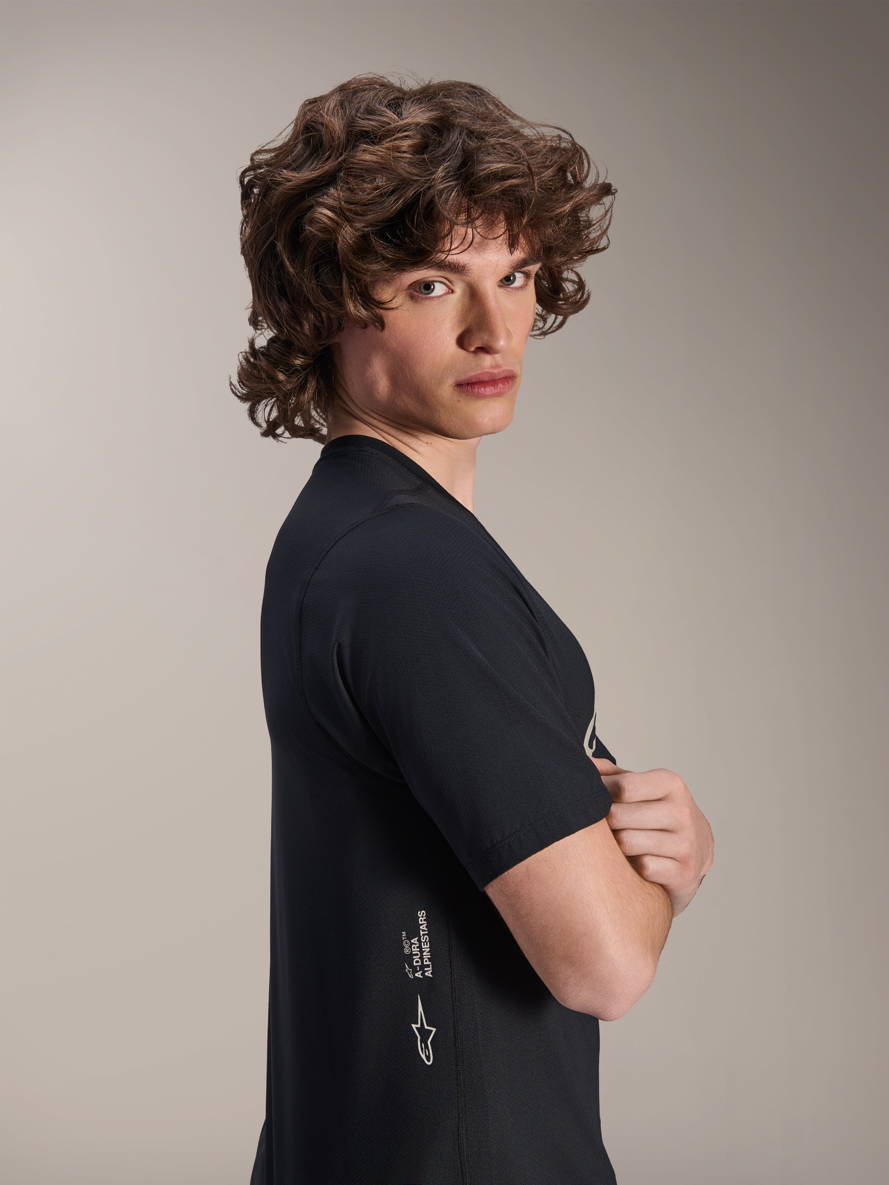 Alpinestars A-Dura Astar Jersey, koszulka MTB z krótkim rękawem, czarna, model pozujący z profilu ze skrzyżowanymi ramionami, białe logo Alpinestars w dolnej części boku, sportowy design do kolarstwa górskiego