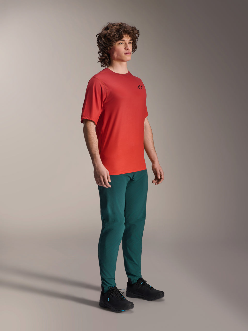 Model ubrany w spodnie Alpinestars A-Dura w kolorze Dark Pine, w połączeniu z czerwoną koszulką z krótkim rękawem A-Dura Rise i czarnymi butami MTB, sportowa odzież do kolarstwa górskiego w studio