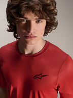 Portret z bliska męskiego modela w koszulce Alpinestars A-Dura Rise z krótkim rękawem w kolorze Spicy Orange, małe czarne logo Astars na klatce piersiowej, wyczynowa odzież MTB