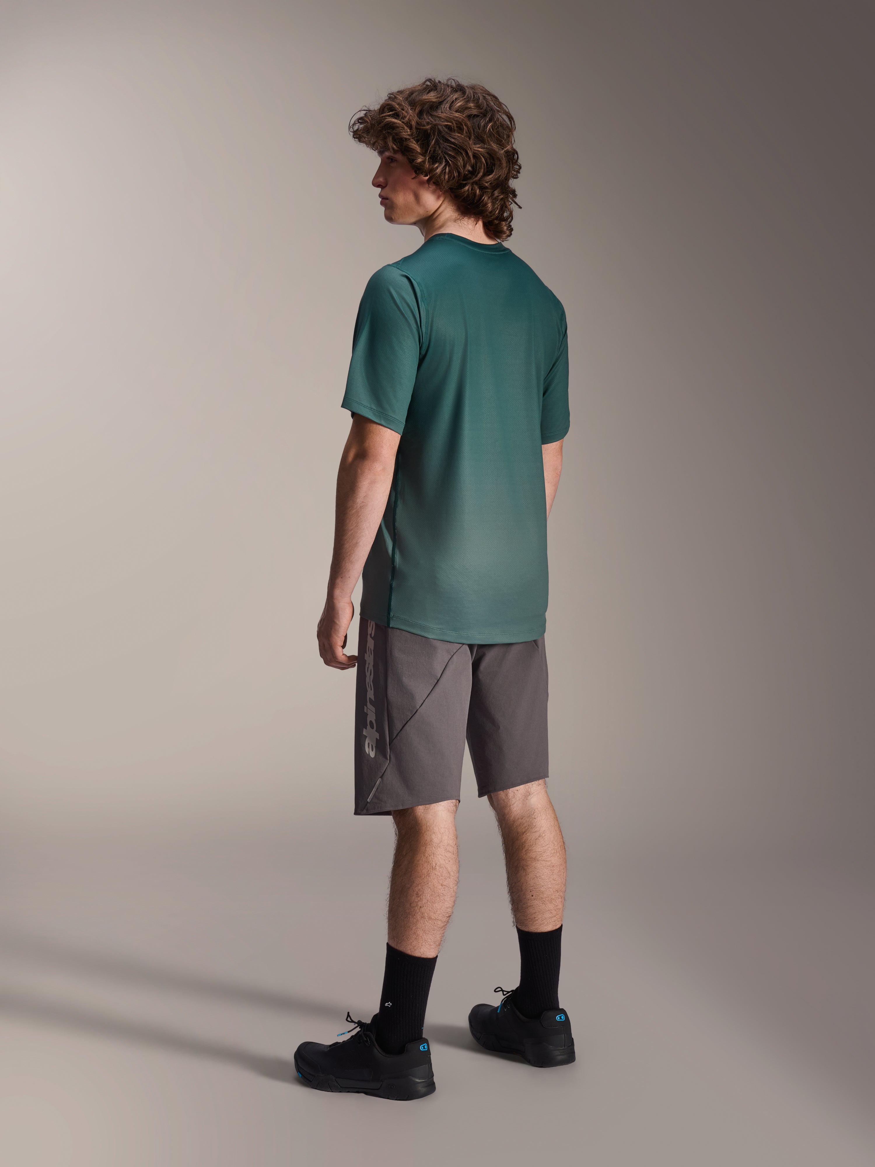 Alpinestars A-Dura Rise Jersey, koszulka MTB z krótkim rękawem, kolor Silver Pine (zielony), widok z tyłu w zestawieniu z szarymi szortami i czarnymi butami, przedłużony tył, regularny krój, lekki materiał odprowadzający wilgoć do jazdy po szlakach