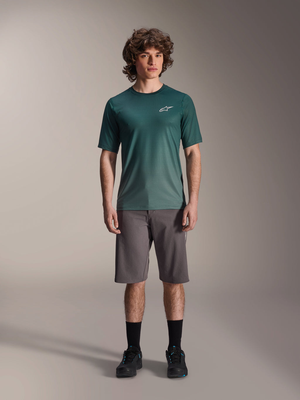 Męski model w koszulce Alpinestars A-Dura Rise Jersey - Short Sleeve w kolorze Silver Pine, zielona koszulka rowerowa z małym białym logo na piersi, zestawiona z szarymi szortami MTB i czarnym obuwiem, swobodny strój na szlaki rowerowe