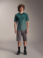 Męski model w koszulce Alpinestars A-Dura Rise Jersey - Short Sleeve w kolorze Silver Pine, zielona koszulka rowerowa z małym białym logo na piersi, zestawiona z szarymi szortami MTB i czarnym obuwiem, swobodny strój na szlaki rowerowe