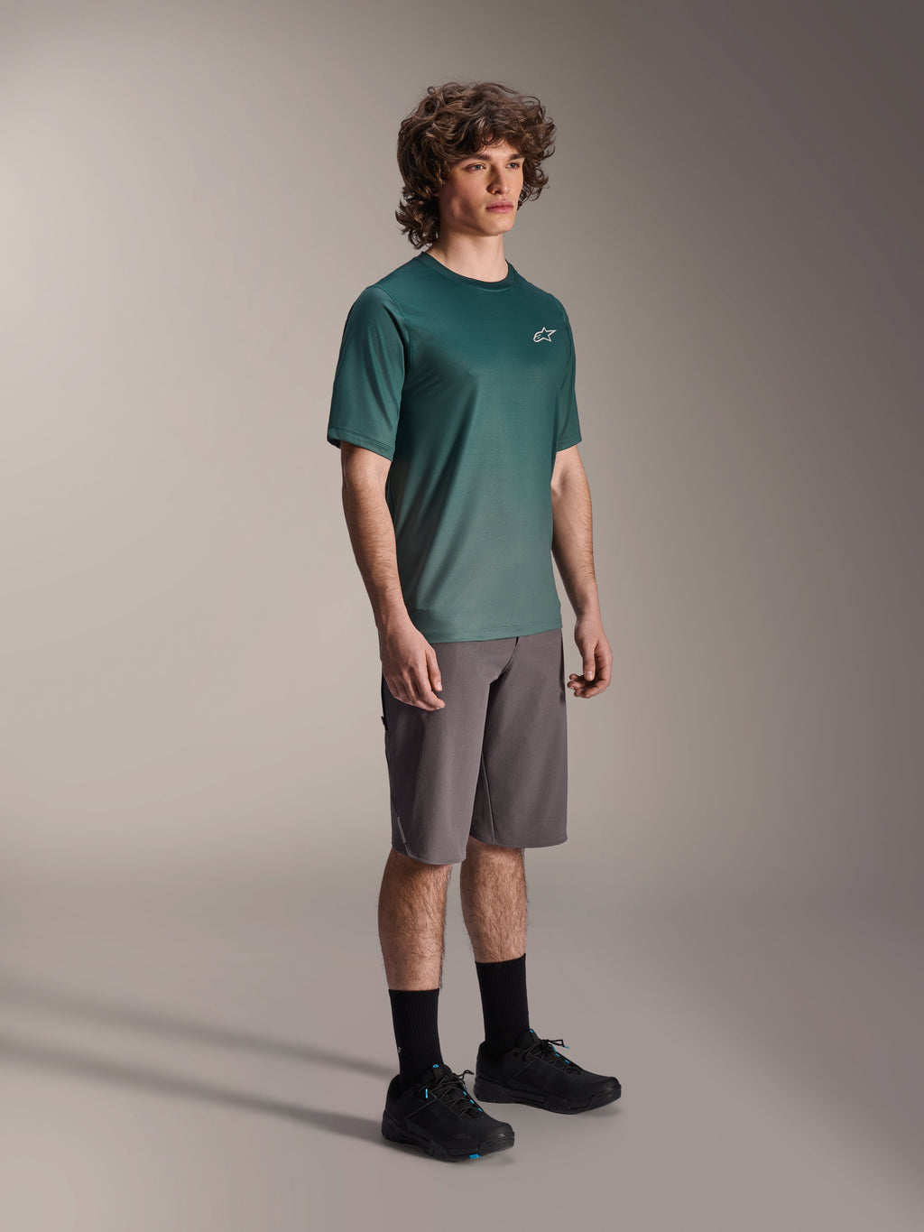Alpinestars A-Dura Rise Jersey - Short Sleeve w kolorze Silver Pine, zielona koszulka rowerowa MTB noszona przez modela z szarymi szortami i czarnymi sneakersami, swobodny strój na szlaki MTB z małym białym logo na klatce piersiowej