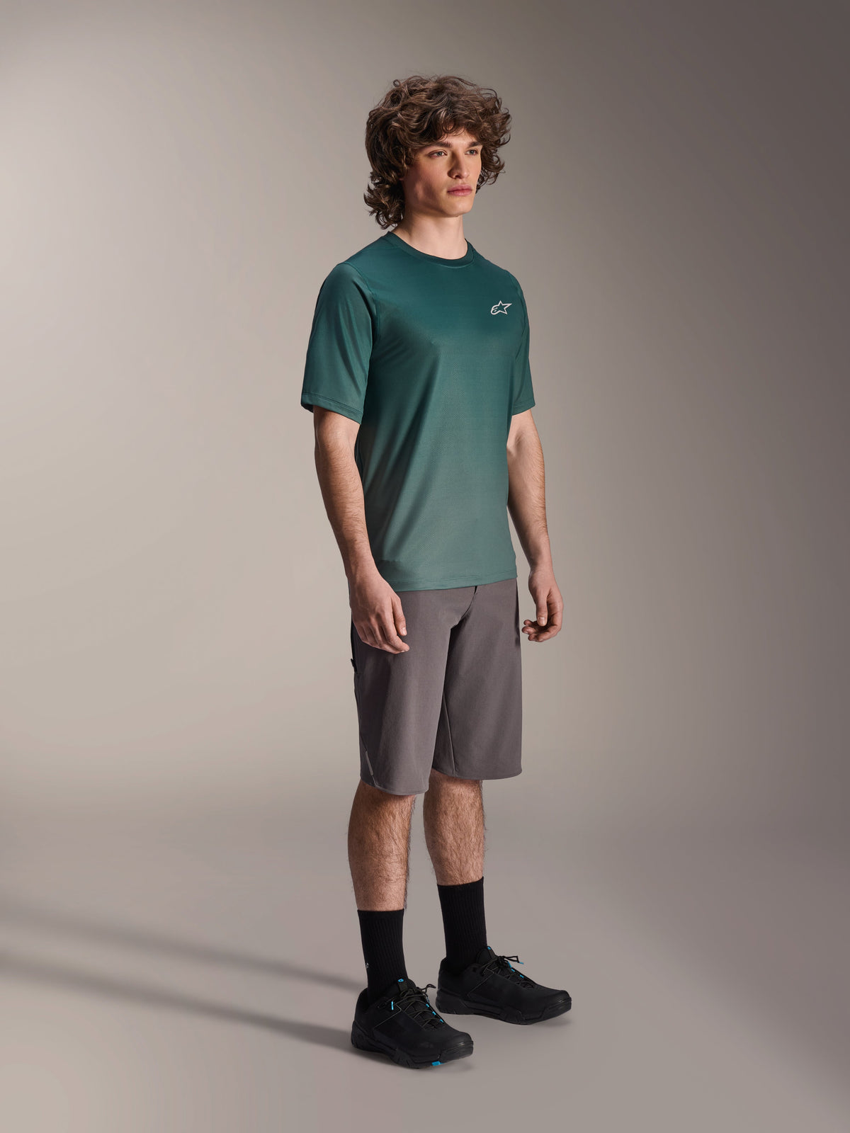 Alpinestars A-Dura Rise Jersey - Short Sleeve w kolorze Silver Pine, zielona koszulka rowerowa MTB noszona przez modela z szarymi szortami i czarnymi sneakersami, swobodny strój na szlaki MTB z małym białym logo na klatce piersiowej