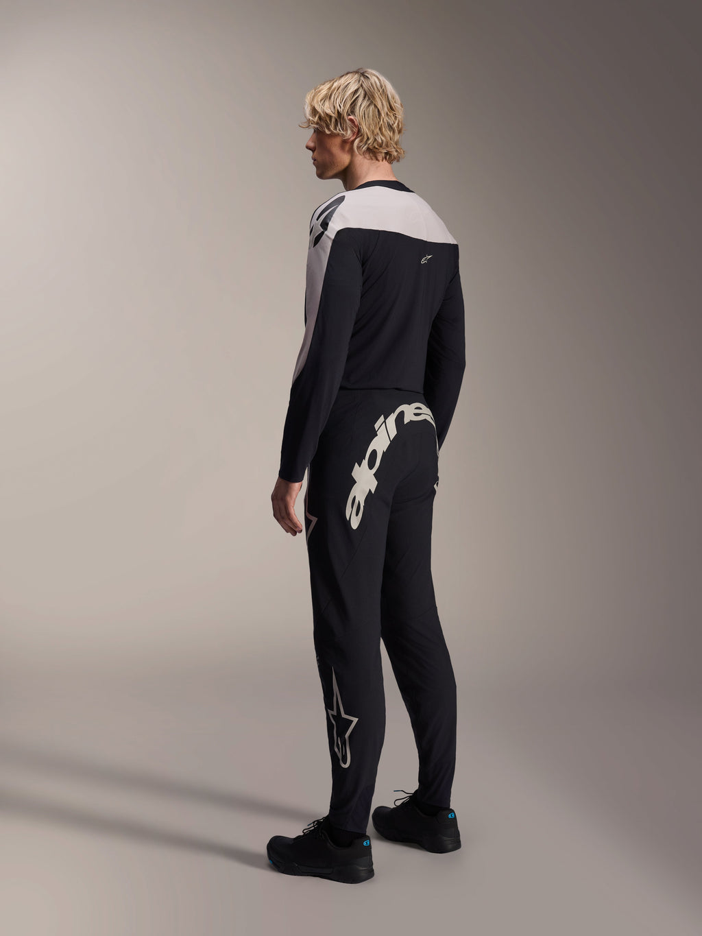 Koszulka Alpinestars A-Supra Melt z długim rękawem, czarna z białymi panelami na ramionach, widok na modela od tyłu i z boku, czarne spodnie MTB z dużym białym napisem, wyczynowy zestaw wyścigowy