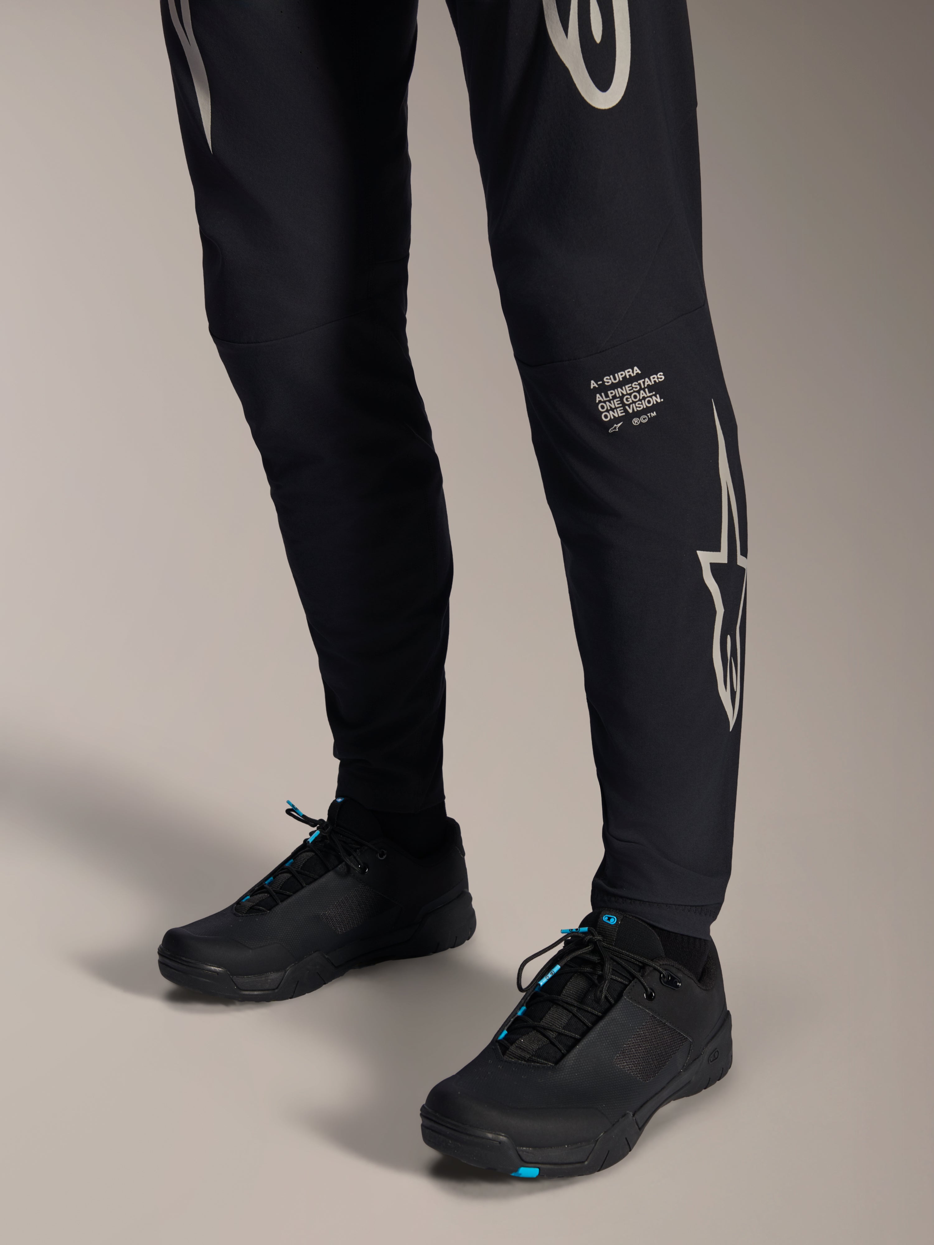 Alpinestars A-Supra Melt Pants, czarne spodnie rowerowe MTB, z białym napisem i logo Alpinestars na nogawce, zestawione z czarnymi butami rowerowymi z niebieskimi akcentami, wyczynowa odzież terenowa