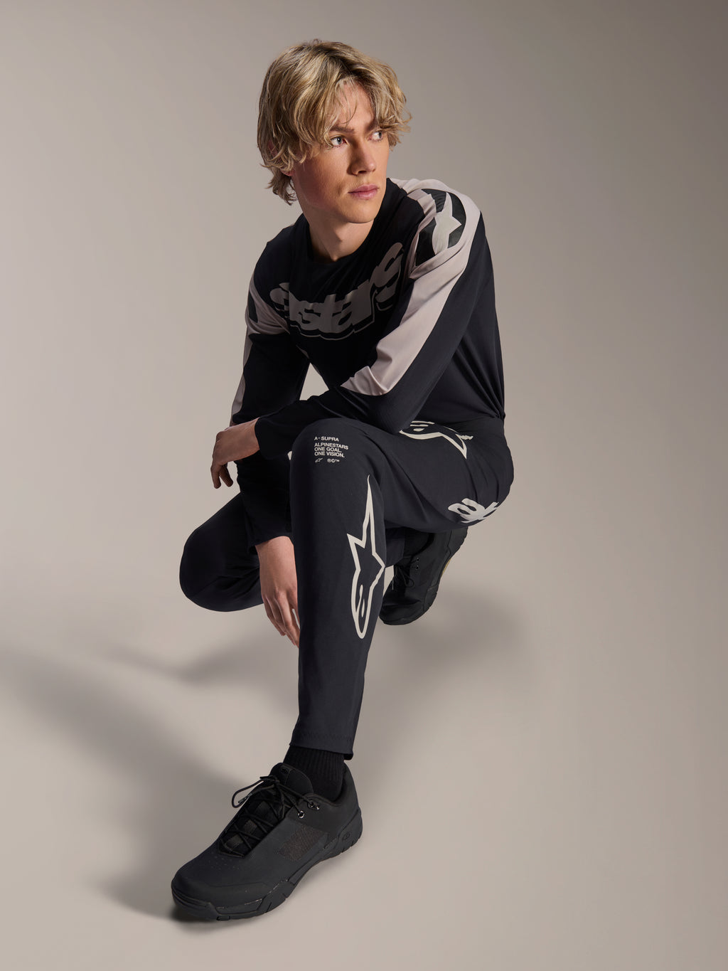 Alpinestars A-Supra Melt Pants i koszulka z długim rękawem, czarno-szare, z wyraźnym białym brandingiem i logo gwiazdy, model w pozycji kucającej, dopasowany zestaw wyścigowy MTB