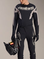 Zestaw wyścigowy MTB Alpinestars A-Supra Melt, koszulka z długim rękawem i spodnie, czarno-szary z białym logo Astars, model trzymający czarny kask full-face i mający na sobie pasujące rękawiczki, wyczynowa odzież do downhillu i enduro