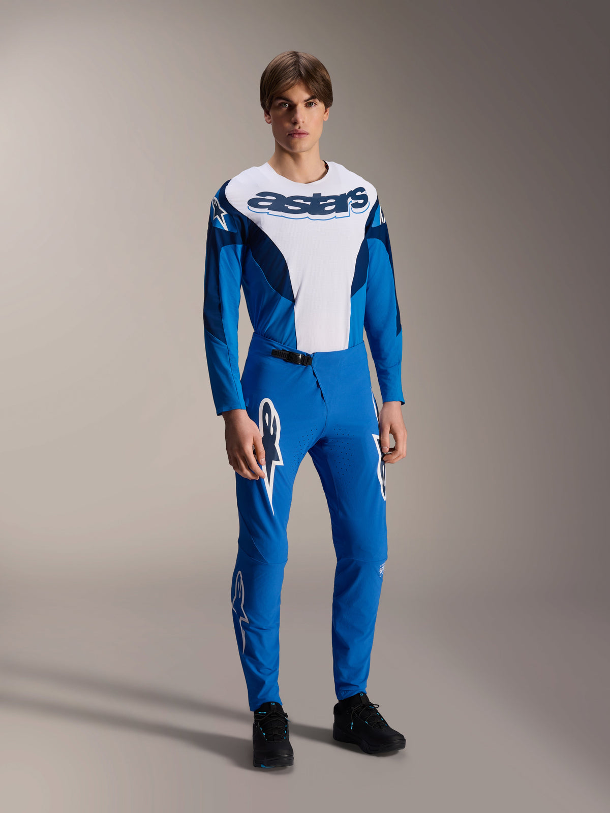 Model w kompletnym stroju wyścigowym MTB Alpinestars, niebiesko-biała koszulka z długim rękawem z brandingiem „astars”, pasujące niebieskie spodnie A-Supra Melt z białymi logotypami gwiazdy, profesjonalna odzież do kolarstwa górskiego do jazdy terenowej