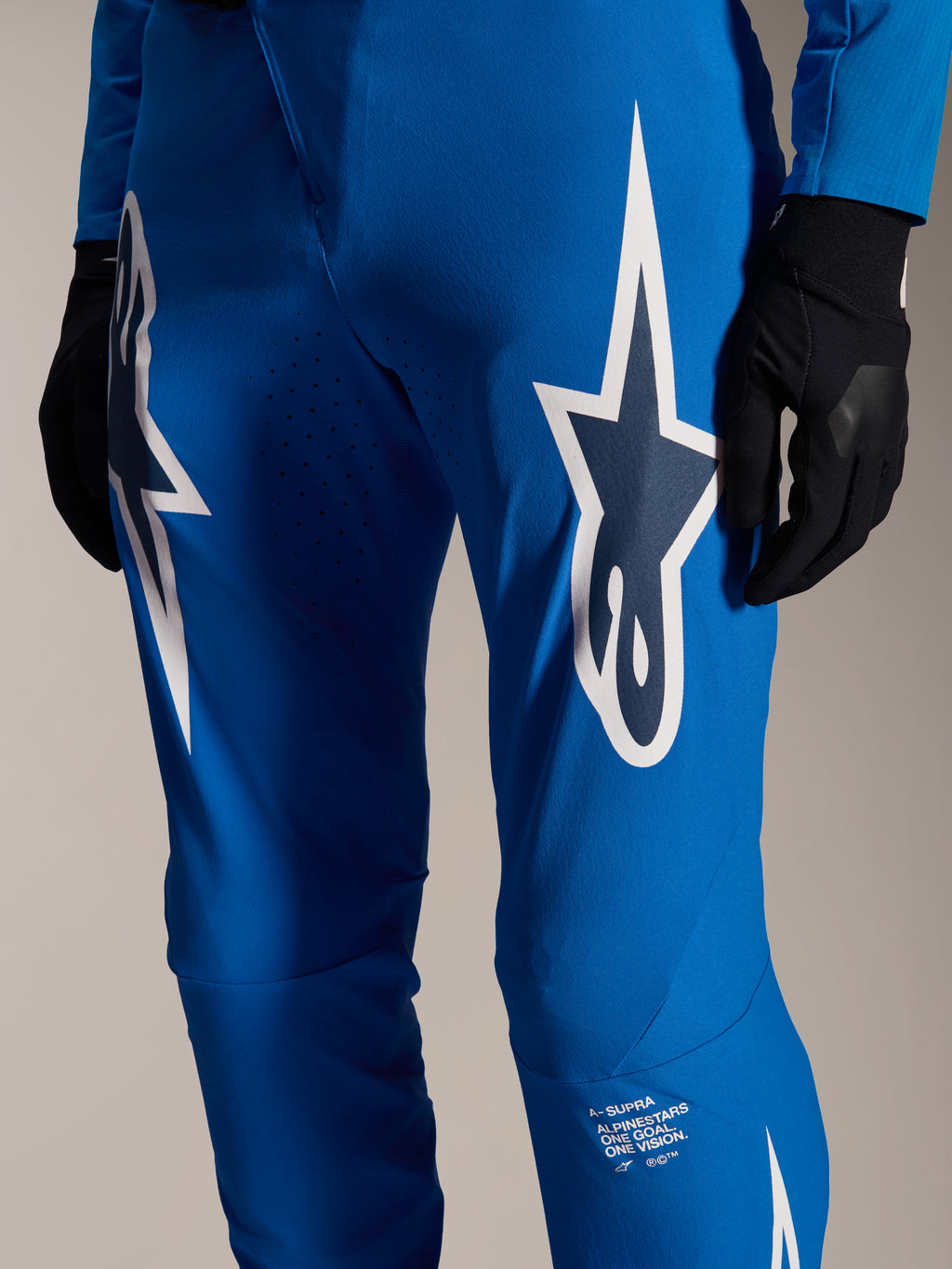 Alpinestars A-Supra Melt Pants, niebieskie spodnie rowerowe, duże białe logo Astars na udach, laserowo wycinane perforacje dla przepływu powietrza, charakterystyczny branding na dole nogawki, czarne rękawiczki, wyczynowa odzież MTB