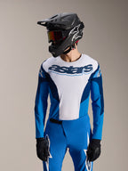 Model ubrany w koszulkę Alpinestars A-Supra Melt z długim rękawem w kolorze niebiesko-białym, z napisem „astars” na klatce piersiowej, w zestawie z dopasowanymi niebieskimi spodniami MTB, czarnym kaskiem typu full-face i goglami; kompletny strój do wyścigów MTB.