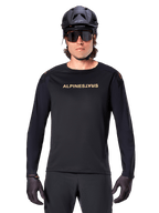Alpinestars A-Aria Polartec Switch Long Sleeve Jersey, czarna, złote logo Alpinestars na piersi, noszona z czarnym kaskiem MTB, ciemnymi okularami i czarnymi rękawiczkami, profesjonalna odzież na rower górski