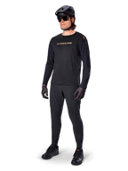 Koszulka z długim rękawem Alpinestars A-Aria Polartec Switch, czarna, brązowe logo na piersi, noszona przez kolarza górskiego w czarnym kasku i okularach przeciwsłonecznych, wydajna odzież rowerowa do jazdy po szlakach