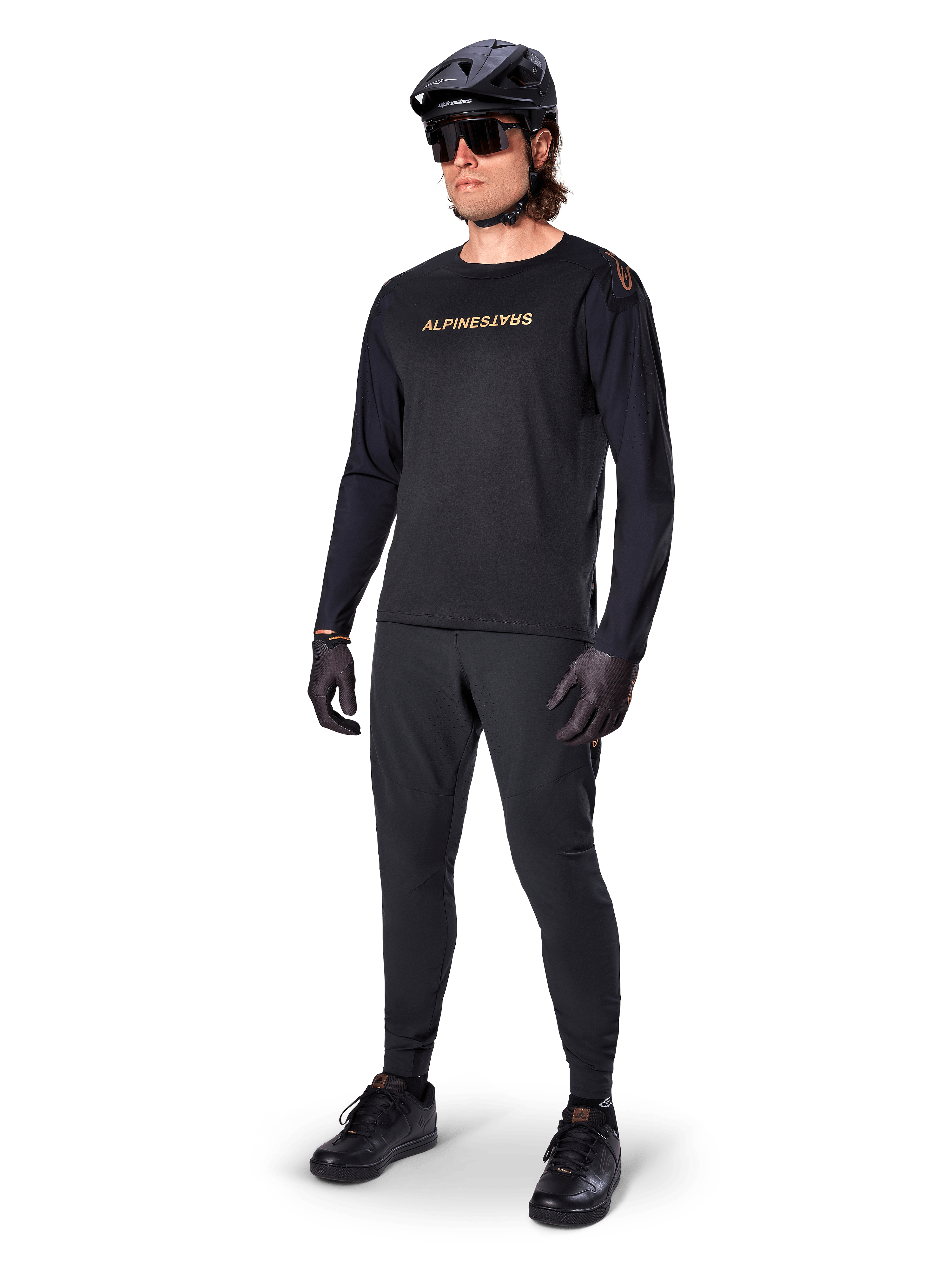 Koszulka z długim rękawem Alpinestars A-Aria Polartec Switch, czarna, brązowe logo na piersi, noszona przez kolarza górskiego w czarnym kasku i okularach przeciwsłonecznych, wydajna odzież rowerowa do jazdy po szlakach