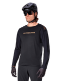 Alpinestars A-Aria Polartec Switch Long Sleeve Jersey, czarna ze złotym logo, odzież do jazdy na rowerze górskim MTB, noszona z czarnym kaskiem i okularami przeciwsłonecznymi, laserowo wycinana wentylacja na rękawach oraz odprowadzający wilgoć materiał Polartec zapewniający wydajność w terenie