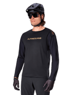 Alpinestars A-Aria Polartec Switch Long Sleeve Jersey, czarna ze złotym logo, odzież do jazdy na rowerze górskim MTB, noszona z czarnym kaskiem i okularami przeciwsłonecznymi, laserowo wycinana wentylacja na rękawach oraz odprowadzający wilgoć materiał Polartec zapewniający wydajność w terenie