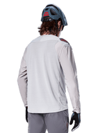 Alpinestars A-Aria Polartec Switch Long Sleeve Jersey, jasnoszara, widok z tyłu na rowerzyście, w zestawieniu z niebieskim kaskiem MTB i szarymi spodniami, techniczna odzież do kolarstwa górskiego na szlaki terenowe